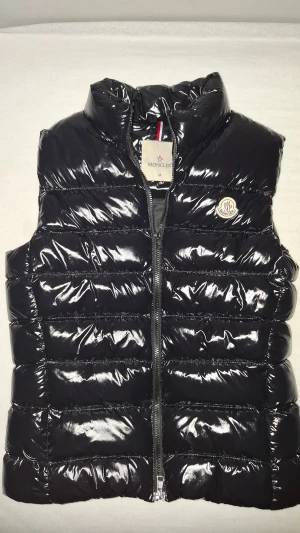 Svart dunväst från Moncler, M - Snygg svart dunväst från Moncler med glansig finish och klassisk logga på bröstet. Västen har quiltad design, hög krage och dragkedja framtill. Insidan har ett coolt serietidningsmönster och etikett med 'Longue Saison'. Perfekt för lager-på-lager och streetstyle.