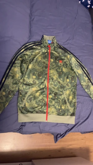 Adidas camo hoodie  - Säljer en grön Adidas hoodie med camo och döskallemönster, röda dragkedjor och klassiska svarta stripes på ärmarna. Stor Adidas-logga på ryggen och mindre logga på bröstet. Jackan har ståkrage och två fickor med dragkedja.