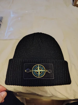 Svart ribbad mössa från Stone Island - Svart ribbstickad mössa från Stone Island med klassisk patch framtill, fäst med två knappar. Märkets logga i blått, gult och grönt ger en clean och exklusiv vibe. Perfekt för dig som vill ha streetstyle med premiumkänsla.