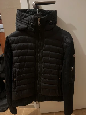 Svart Moncler pufferjacka med huva - Svart pufferjacka från Moncler med stickade ärmar och ribbade muddar. Jackan har huva med snörning, dragkedja framtill och Moncler-logga på ärmen. Materialmix av polyester och stickad textil, perfekt för kyliga dagar. Snygg, stilren och riktigt skön att bära.