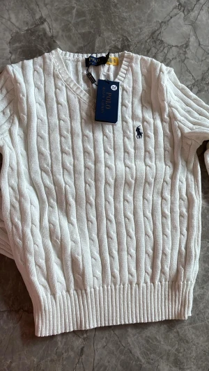 Vit kabelstickad tröja Polo Ralph Lauren - Säljer en vit kabelstickad tröja från Polo Ralph Lauren med v-ringad hals och broderad logga på bröstet. Tröjan har långa ärmar och ribbade muddar vid ärmslut och nederkant. Perfekt för dig som gillar klassisk och stilren look.