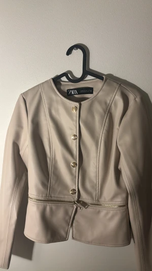 Beige skinnjacka från Zara XS - Snygg beige skinnjacka från Zara i storlek XS. Jackan har rund hals, guldfärgade knappar framtill och en dekorativ dragkedja vid midjan. Stilren design med långa ärmar och figurnära passform. Perfekt för dig som gillar minimalistisk och chic look.