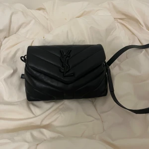 Svart quiltad axelväska från Saint Laurent - Snygg svart axelväska från Saint Laurent med ikoniskt YSL-emblem framtill. Väskan är quiltad i ett V-mönster och har justerbar axelrem. Tillverkad i mjukt skinn med magnetlås och innerfack med dragkedja. Perfekt storlek för det viktigaste. Inte fast pris.