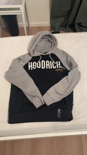 Svart och grå hoodie från Hoodrich S - Svart och grå hoodie från Hoodrich i storlek S. Tröjan har huva med snörning, kontrastfärgade ärmar och tryck i vitt och gult både fram och bak. Materialet är mjuk bomull och polyester, perfekt för chill dagar. Snyggt broderad FNTS-detalj på ärmen.