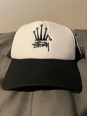 Stüssy svartvit truckerkeps - Snygg truckerkeps från Stüssy i svart och vitt med klassisk Stüssy-logga och kronbroderi framtill. Kepsen har mesh på sidorna och justerbar snapback baktill. Perfekt för streetwear och att lyfta din stil.