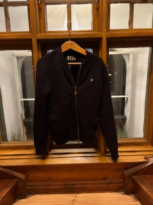 Marinblå tröja från Morris - Marinblå tröja från Morris i storlek S. Tröjan har en stilren design med ribbade muddar dragkedja framtill och ett litet broderat emblem på bröstet. Den är i mycket bra skick