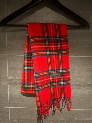 Rutig röd halsduk från Burberrys - Snygg rutig halsduk från Burberrys i klassisk röd färg med inslag av blått, gult, grönt och vitt. Gjord i 100% lammull och har fransar i ändarna. Perfekt för att ge din outfit en cool brittisk vibe.