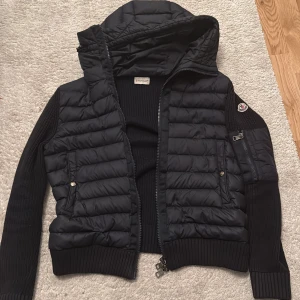 Mörk blå moncler cardigan jacka  från Moncler - Jag säljer en äkta mörk blå  Moncler cardigan  med stickade ärmar och quiltad kropp. Jackan har huva, dragkedja framtill och på ärmen, samt ribbade muddar. Snygg Moncler-logga på axeln och praktiska fickor med knappar. Perfekt för dig som gillar stilren och sportig look. Storlek S och är i bra skick 