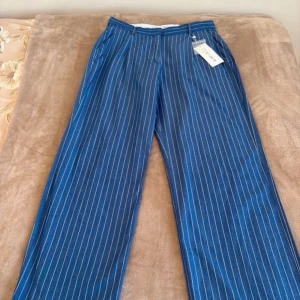 Blå randiga wide byxor från Amisu - Snygga blå byxor från Amisu med vita vertikala ränder och breda ben. Byxorna har normal passform, midwaist och klassisk knappstängning. Perfekta för dig som gillar en trendig och avslappnad look. Materialet känns lätt och bekvämt.