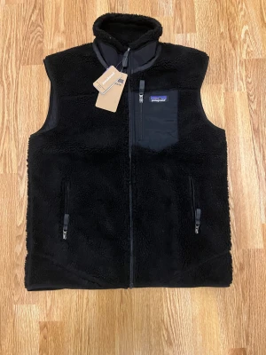 Svart fleeceväst från Patagonia - Svart fleeceväst från Patagonia med hög krage och hel dragkedja framtill. Västen har två sidofickor med dragkedja och en bröstficka med dragkedja och logotyp. Materialet är mjuk fleece som ger en skön och varm känsla. Perfekt för lager-på-lager under kyliga dagar.