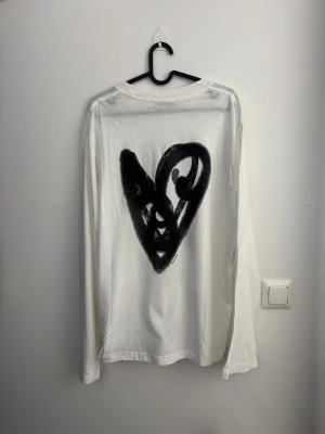 Vit långärmad tröja Acne Studios med hjärta i velour storlek M - Vit långärmad tröja från Acne Studios i mjuk bomull. Stor svart hjärtdetalj med grafiskt mönster och i velour på ryggen och mindre hjärta framtill. Rund hals och relaxed passform, perfekt för dig som gillar stilrena plagg med en twist.