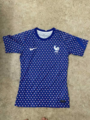 Blå prickig Frankrike fotbollströja Nike - Snygg blå fotbollströja från Nike med Frankrikes landslagsmärke på bröstet. Tröjan har korta ärmar och är täckt av ett prickigt mönster i vitt och rött. Tillverkad i lätt och ventilerande polyester, perfekt för match eller träning.