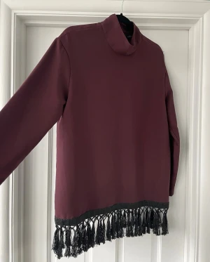Vinröd topp med fransar från Zara - Säljer en vinröd topp från Zara Woman i storlek S. Toppen har lång ärm, hög krage och  svarta fransdetaljer längst ner. Materialet är mjukt och känns som polyester. Dragkedja i ryggen för enkel påklädning. 