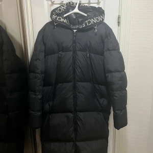 Svart Moncler lång pufferjacka - Kom privat för prisförslag Svart lång pufferjacka från Moncler med stor huva och logotyptryck längs kanten. Jackan har två dragkedjefickor framtill, quiltad design och Moncler-märke på ärmen. Perfekt för kalla dagar med sin rejäla fyllning och stilrena look.  