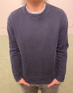 Mörkblå sweatshirt från J.Lindeberg - Snygg mörkblå sweatshirt från J.Lindeberg med diskret broderad logga på bröstet. Tröjan har rund hals, ribbade muddar och är tillverkad i mjuk bomull. Perfekt för dig som gillar stilrena och bekväma plagg med en clean vibe.