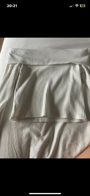 Vit ribbad offshoulder topp - Säljer en vit ribbad topp med offshoulder-design och långa ärmar. Toppen har en bred kant upptill som ger en snygg och modern look. Perfekt för dig som gillar stilrena och trendiga plagg med lite edge.