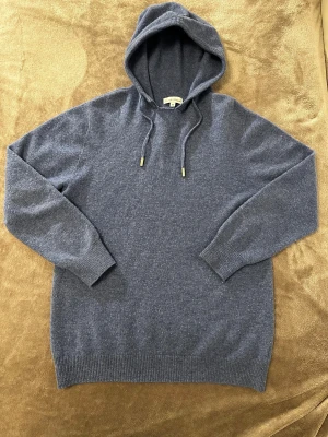 Blå hoodie i merinoull från GardeGrisch - Snygg mörkblå hoodie från GardeGrisch i storlek XS, tillverkad i 100% mjuk merinoull. Tröjan har huva med snörning och guldfärgade detaljer på snörets ändar. Långärmad modell med ribbade muddar och klassisk loose passform. Perfekt för kyliga dagar. liten defekt se bild 3 NYPRIS 979kr