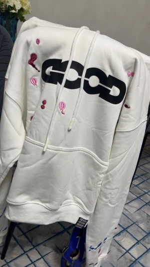 Vit hoodie från Good med broderi - Vit hoodie från Good med stor svart logga på bröstet och broderade detaljer i rosa och rött. Tröjan har huva med snörning och en klassisk magficka. Materialet är mjuk bomull och passformen är normal. Perfekt för dig som gillar streetwear med unika detaljer.