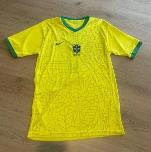 Brasilien Nike fotbollströja - Gul Brasilien fotbollströja med gröna detaljer från Nike, Vini Jr. och nummer 7 på ryggen. Tröjan har korta ärmar, V-ringning med grönt kantband och ett grafiskt mönster över hela framsidan. Broderad Nike-logga och CBF-emblem på bröstet. Tillverkad i lätt polyester.
