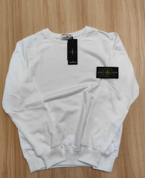 Vit sweatshirt från Stone Island - Säljer en stilren vit sweatshirt från Stone Island med klassisk rund hals och ribbade muddar. Tröjan har den ikoniska Stone Island-loggan på vänster ärm och är tillverkad i mjuk bomull. Perfekt för dig som gillar clean och enkel streetwear.