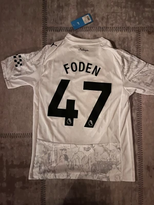Manchester City Foden #47 Puma tröja - Säljer en officiell Manchester City fotbollströja från Puma med Foden #47 på ryggen. Tröjan är vit med grått grafiskt mönster, har korta ärmar och en diagonal röd och svart rand på framsidan. Materialet är lätt polyester med RE:FIBRE-teknologi. Perfekt för dig som älskar City!