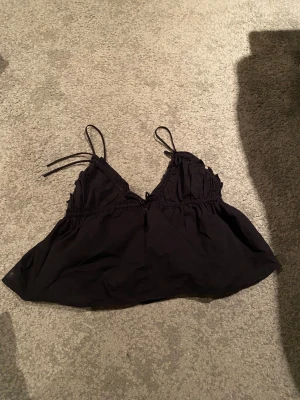 Svart linnetopp med volangdetaljer - Säljer en svart linnetopp med tunna axelband och volangdetaljer längs bysten. Toppen har en lätt rynkad och luftig passform, samt en söt knytning framtill. Den går även ner i ryggen och har en söt rosset där bak, perfekt för alla tillfällen🥰