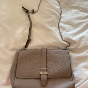 Beige axelväska från Anna Field - Snygg beige axelväska från Anna Field med kedjerem och lock i reptilmönstrad skinnimitation. Väskan har ett stilrent metallspänne framtill och flera fack inuti, inklusive ett med dragkedja. Perfekt storlek för det viktigaste och passar till många outfits.