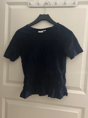 Mörkblå kortärmad topp - Säljer en mörkblå topp från gina med korta ärmar och rund hals. Toppen har en lätt rynkad struktur och vågig kant nedtill som ger en cool och unik look. Perfekt att styla med jeans eller kjol för en chill vibe.