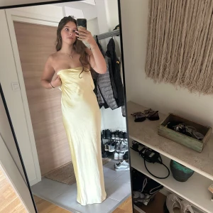 Gul långklänning i satin - Säljer en elegant balklänning i gul satin. Klänningen är axelbandslös med en snygg öppen rygg och har en figurnära passform som faller mjukt längs kroppen. Perfekt för bal 😍 klänningen har två revor som syns på de sista bilderna annars är den som i ny skick😍