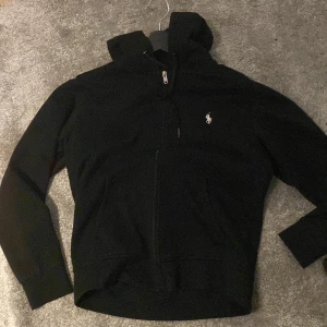 Svart hoodiejacka från Polo Ralph Lauren - Svart hoodiejacka från Polo Ralph Lauren med dragkedja framtill och klassisk logga på bröstet. Jackan har huva, långa ärmar och två fickor framtill. Perfekt för chill dagar och enkel att matcha med jeans eller joggers.
