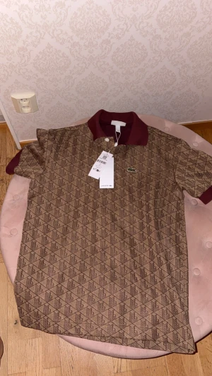 Brun mönstrad piké från Lacoste - Snygg brun pikétröja från Lacoste med geometriskt mönster och klassisk krokodil-logga på bröstet. Krage och ärmslut i vinröd ton ger en schysst kontrast. Perfekt för dig som vill sticka ut med stilren design och coola detaljer.