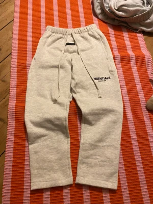 Essentials Fear of God mjukisbyxor - Snygga ljusgrå mjukisbyxor från Essentials Fear of God med svart logga på benet. Byxorna har elastisk midja med snörning och raka ben. Tillverkade i mjukt bomullsmaterial som är perfekt för chill eller träning. Klassisk och clean look.
