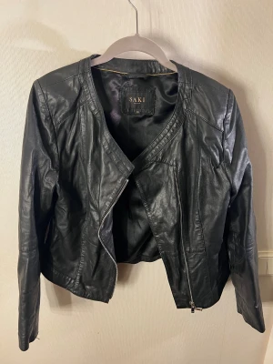 Svart skinnjacka från Saki - Svart skinnjacka från Saki med snygg, stilren design och rundad hals. Jackan har något diagonal dragkedja framtill och är figursydd med coola sömmar. Tillverkad i äkta skinn med polyesterfoder. Använd men inga anmärkningar.