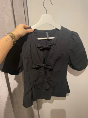 Svart blus med rosetter från Stradivarius  - Supersöt svart blus från Stradivarius med puffiga korta ärmar och tre knytrosetter framtill. Blusen har en lätt v-ringad hals och volangkant nedtill som ger en feminin vibe❣️