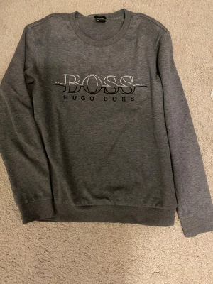 Grå slim fit tröja från Hugo Boss - Snygg grå långärmad tröja från Hugo Boss med stort BOSS-tryck på bröstet. Slim fit-modell med rund hals och ribbade muddar. Perfekt för dig som gillar stilrena och klassiska plagg med en modern touch.