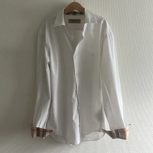 Äkta Burberry Skjorta - Säljer en klassisk vit Burberry skjorta. Den har en krage med rutigt mönster inuti och om man viker upp ärmarna på den så är det ett rutigt mönster inuti.