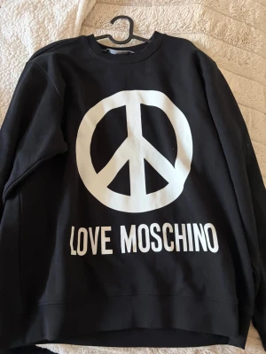 Svart sweatshirt Love Moschino XL - Svart sweatshirt från Love Moschino i storlek XL med stort vitt peace-märke och text på framsidan. Tröjan har rund hals, lång ärm och är gjord i mjuk bomull. Perfekt statement-plagg för dig som gillar streetstyle och tydliga prints.