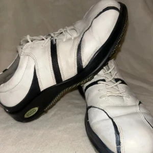 Vita och svarta sneakers från Ecco - Snygga sneakers från Ecco i vitt skinn med svarta detaljer och snörning. Skorna har en sportig look med rund tå och platt sula, perfekta för en avslappnad stil. Klassisk design med kontrasterande svarta ränder på sidorna och svart sula.  Perfekt för golfrundan! Storlek 37