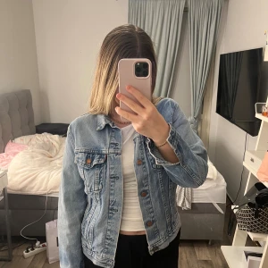 Jeansjacka Levi's - Säljer en klassisk ljusblå jeansjacka från Levi's med snygga bronsfärgade knappar och två bröstfickor med lock. Jackan har en rak passform och är tillverkad i slitstarkt denim. Perfekt att slänga över en t-shirt för en chill look.