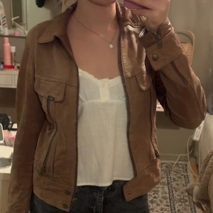Brun skinnjacka från Lee, storlek S - Säljer en snygg brun skinnjacka från Lee i storlek S. Jackan har klassisk krage, flera fickor med dragkedja och knappdetaljer vid ärmslut. Passformen är normal och jackan har en stilren look som funkar till jeans och t-shirt.