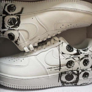 Nike Air Force 1 x Comme des Garçons Supreme - Säljer ett par vita Nike Air Force 1 i collab med Comme des Garçons och Supreme. Skorna har unikt svart ögonmönster på sidorna och textdetalj. Klassisk siluett med snörning och perforerad tå. Perfekta för dig som vill sticka ut med streetwear-vibe.