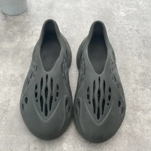 Adidas Yeezy Foam Runner svart - Svarta Adidas Yeezy Foam Runner med futuristisk design och många ventilerande hål. Tillverkade i lätt syntetmaterial som ger en skön och luftig känsla. Perfekta för dig som vill sticka ut med en unik och modern look.