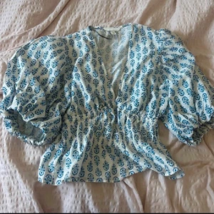 Blommig blus med puffärm - Säljer en söt blus med blått blommigt mönster på vit botten. Blusen har puffiga korta ärmar, v-ringad hals och smockad midja som ger en snygg siluett. Perfekt för dig som gillar romantisk stil och vill sticka ut lite extra.