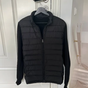 Svart cardigan med dragkedja - Svart cardigan med quiltad front och släta stickade ärmar. Jackan har hel dragkedja framtill, ståkrage och två sidofickor med dragkedja. Perfekt för lager-på-lager och enkel att matcha med olika outfits. Denna cardigan är perfekt för hösten!