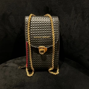 Svart axelväska från Laura Ashley - Säljer en svart axelväska från Laura Ashley med 3D-mönstrad yta och guldfärgad kedja. Väskan har ett elegant lås framtill och insidan är klarröd med flera fack och dragkedja. Perfekt för mobilen och småprylar när du vill ha något stilrent och unikt.