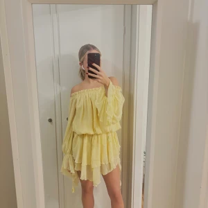 Gul offshoulder kortklänning - Superfin ljusgul kortklänning med offshoulder och volangdetaljer. Klänningen har långa, vida ärmar och flera lager i kjolen som ger en flowy känsla. .