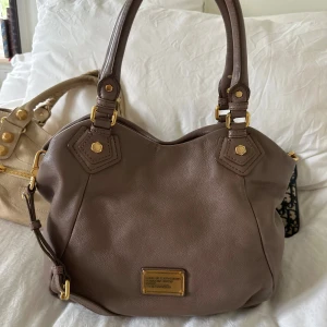 Brun Q Classic Fransesca Bag! - Rymlig och stilren Marc by Marc Jacobs Classic Q Francesca i mjukt beige brunt pebble-läder med guldfärgade detaljer. En klassiker från den ikoniska Classic Q-serien som är både praktisk och tidlös. Perfekt för dig som vill ha en snygg vardagsväska där även en 13” MacBook får plats utan problem.  Väskan har dubbla handtag och en avtagbar axelrem, så den kan bäras både i handen och över axeln. Invändigt finns flera fack och dragkedja för att hålla ordning på dina saker.  Mått: ungefär 40×30×10 cm