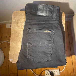 Svarta jeans från Nudie Jeans - Svarta jeans från Nudie Jeans med klassisk femficksdesign och subtil broderad våg på bakfickan. Jeansen har en svart läderpatch i midjan och är tillverkade i mjukt bomullsmaterial. Perfekt för dig som gillar stilrena och tidlösa jeans.