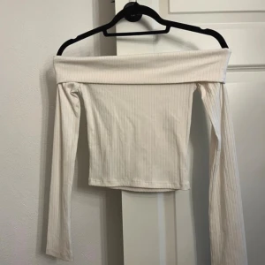 Offshoulder ribbad topp i ljus beige - Trendig offshoulder topp i ljus beige med ribbad struktur och långa ärmar. Perfekt för dig som gillar en stilren och enkel look. Toppen är croppad och har en smickrande passform som framhäver axlarna.