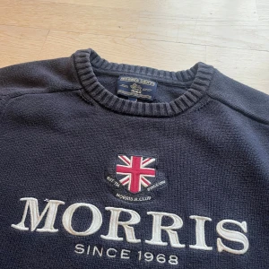 Mörkblå stickad tröja från Morris - Mörkblå stickad tröja från Morris med broderad Union Jack och stor vit logga framtill. Klassisk rund halsringning och ribbade muddar vid ärmslut och nederkant. Perfekt för dig som gillar preppy stil och snygga detaljer.